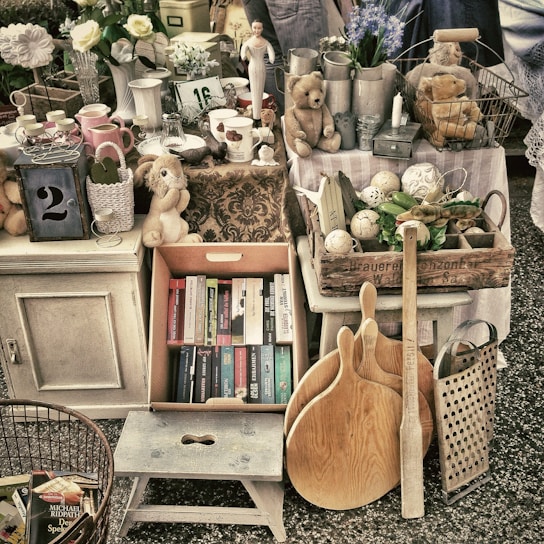 A collection of vintage and antique items arranged in a marketplace setting. The assortment includes books, plush toys, wooden kitchen utensils, ceramic pitchers, a lantern with the number 2, various kitchenware, and decorative objects such as a small statue and flowers. The colors are muted with a rustic and nostalgic feel.