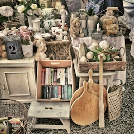A collection of vintage and antique items arranged in a marketplace setting. The assortment includes books, plush toys, wooden kitchen utensils, ceramic pitchers, a lantern with the number 2, various kitchenware, and decorative objects such as a small statue and flowers. The colors are muted with a rustic and nostalgic feel.