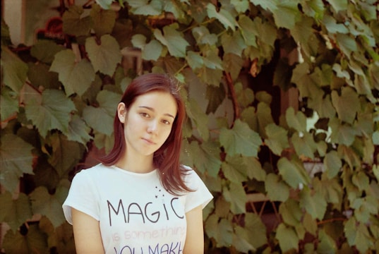 A woman with long brown hair is wearing a white T-shirt with an inspirational message. Behind her, a dense wall of green leafy vines covers a structure, creating a natural backdrop.
