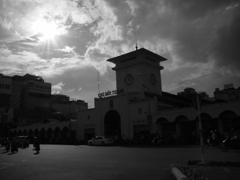 A historic market building is silhouetted against a cloudy sky with the sun peeking through, casting dramatic light and shadows. The clock tower stands prominently above, with vehicles and people seen in the foreground, suggesting a bustling urban environment.