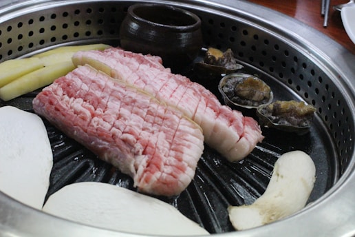 A sizzling grill filled with raw slices of pork belly meat, mushrooms, and shellfish. The food items are arranged neatly on a circular grill, with a small brown ceramic pot in the background. The pinkish hue of the pork contrasts with the earthy tones of the mushrooms and shellfish.