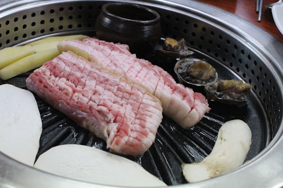 A sizzling grill filled with raw slices of pork belly meat, mushrooms, and shellfish. The food items are arranged neatly on a circular grill, with a small brown ceramic pot in the background. The pinkish hue of the pork contrasts with the earthy tones of the mushrooms and shellfish.
