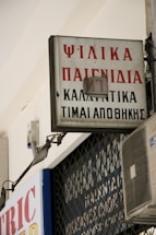 A rectangular sign with Greek text is mounted on a building, displaying red and black lettering. An air conditioning unit is visible below the sign, attached to the wall. The sign appears slightly weathered and is part of an urban environment with metal bars in the background.