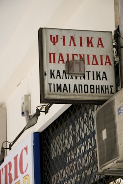 A rectangular sign with Greek text is mounted on a building, displaying red and black lettering. An air conditioning unit is visible below the sign, attached to the wall. The sign appears slightly weathered and is part of an urban environment with metal bars in the background.
