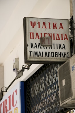 A rectangular sign with Greek text is mounted on a building, displaying red and black lettering. An air conditioning unit is visible below the sign, attached to the wall. The sign appears slightly weathered and is part of an urban environment with metal bars in the background.