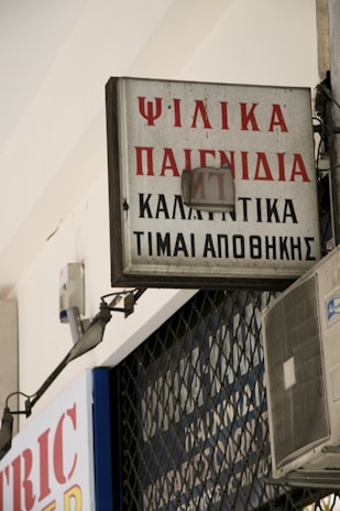 A rectangular sign with Greek text is mounted on a building, displaying red and black lettering. An air conditioning unit is visible below the sign, attached to the wall. The sign appears slightly weathered and is part of an urban environment with metal bars in the background.
