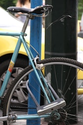 A vintage bicycle with a light blue frame is securely locked to a blue metal post. The focus is on the rear wheel and seat area, showing clear details of the tire, spokes, and lock. The background features a blurred yellow car and parts of another vehicle, along with greenery and a passerby.