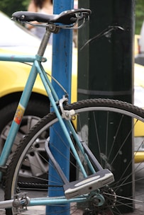 A vintage bicycle with a light blue frame is securely locked to a blue metal post. The focus is on the rear wheel and seat area, showing clear details of the tire, spokes, and lock. The background features a blurred yellow car and parts of another vehicle, along with greenery and a passerby.