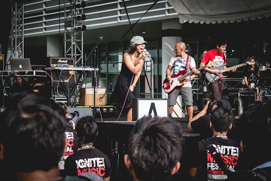 A group of musicians performs on an outdoor stage with a singer in a black dress and hat in the center, accompanied by a bassist and a guitarist. Several amplifiers and electronic equipment are set up, with a small crowd watching in the foreground. The event appears to be a music festival.