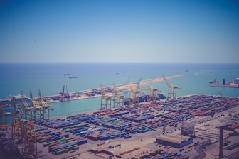 Aerial view of a large shipping port with numerous colorful shipping containers neatly arranged on the docks. Several large cranes are positioned throughout the port for loading and unloading cargo. The clear, blue sea stretches into the background, with a few cargo ships visible on the water. The sky is bright and clear, complementing the tranquil seascape.