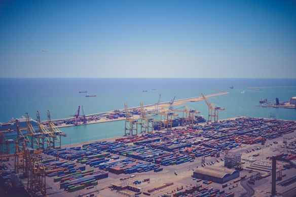 Aerial view of a large shipping port with numerous colorful shipping containers neatly arranged on the docks. Several large cranes are positioned throughout the port for loading and unloading cargo. The clear, blue sea stretches into the background, with a few cargo ships visible on the water. The sky is bright and clear, complementing the tranquil seascape.