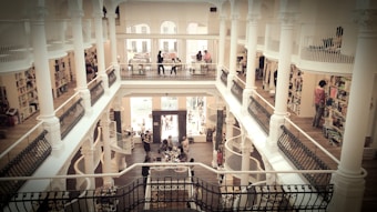A multi-level, ornate library or bookstore featuring white columns, balustrades, and decorative railings. People are browsing the shelves on different levels, and there are spiral staircases connecting the floors. The space is bright with natural light coming from large windows.