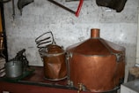 A vintage distillery setup with large copper tanks and coiled pipes is arranged against a scratched and worn wall. Various old tools and metal objects, including a saw and pump, are scattered around.