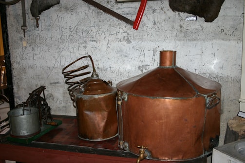 A vintage distillery setup with large copper tanks and coiled pipes is arranged against a scratched and worn wall. Various old tools and metal objects, including a saw and pump, are scattered around.