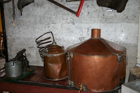 A vintage distillery setup with large copper tanks and coiled pipes is arranged against a scratched and worn wall. Various old tools and metal objects, including a saw and pump, are scattered around.