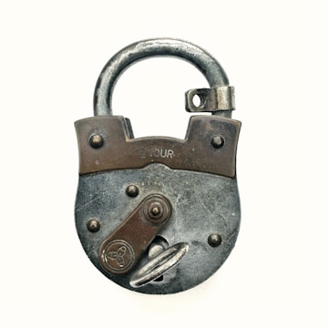 An old, rustic padlock with a round body and a curved shackle. The lock has a worn appearance with visible metallic and rusty textures. It features a lever and a circular emblem in its design.