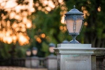 An ornate outdoor lantern sits atop a stone pillar, with the background softly blurred. The scene captures the warm glow of a sunset filtering through the leaves of nearby trees, creating a tranquil ambiance.