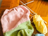 A knitted piece with pink and green stripes lies on a wooden table. A ball of yellow yarn is nearby, with knitting needles inserted into the yarn.