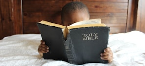 boy reading Holy Bible while lying on bed