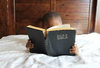 boy reading Holy Bible while lying on bed