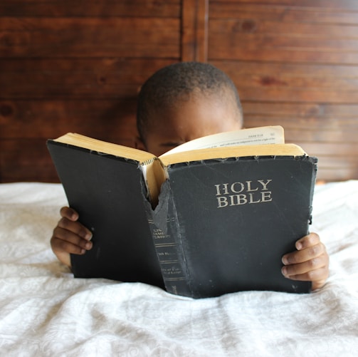 boy reading Holy Bible while lying on bed