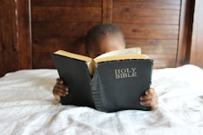boy reading Holy Bible while lying on bed