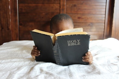 boy reading Holy Bible while lying on bed