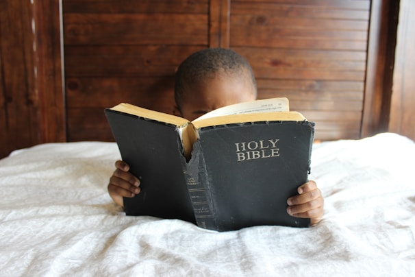 A child is reading a large, worn copy of the Holy Bible while lying on a bed. The wooden headboard is visible in the background, and the child holds the book with both hands. The cover of the Bible is black with white lettering.