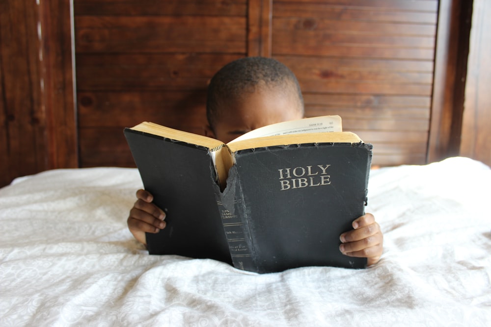 boy reading Holy Bible while lying on bed