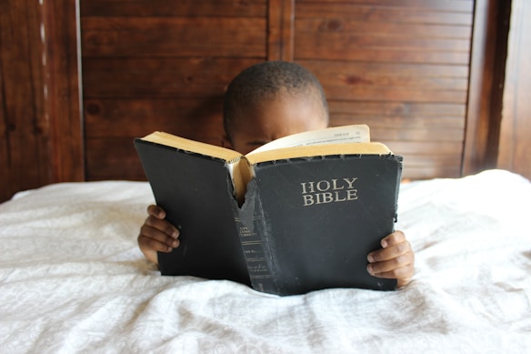 A child is reading a large, worn copy of the Holy Bible while lying on a bed. The wooden headboard is visible in the background, and the child holds the book with both hands. The cover of the Bible is black with white lettering.