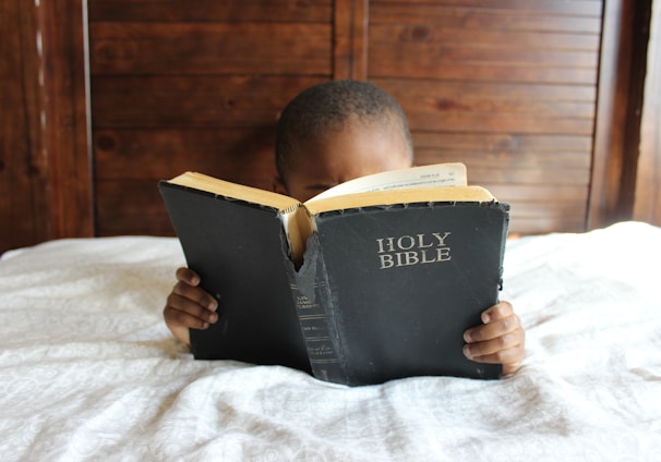 boy reading Holy Bible while lying on bed