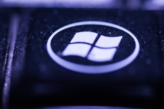 A close-up view of a key on a keyboard featuring a recognizable Windows logo. The key is predominantly black with the logo in white, encircled within a glossy surface. The lighting creates a moody, bluish-purple hue over the key, highlighting its textured surface.