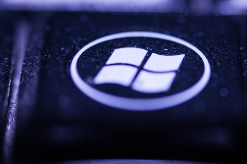 A close-up view of a key on a keyboard featuring a recognizable Windows logo. The key is predominantly black with the logo in white, encircled within a glossy surface. The lighting creates a moody, bluish-purple hue over the key, highlighting its textured surface.