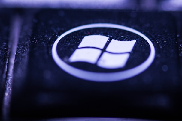 A close-up view of a key on a keyboard featuring a recognizable Windows logo. The key is predominantly black with the logo in white, encircled within a glossy surface. The lighting creates a moody, bluish-purple hue over the key, highlighting its textured surface.