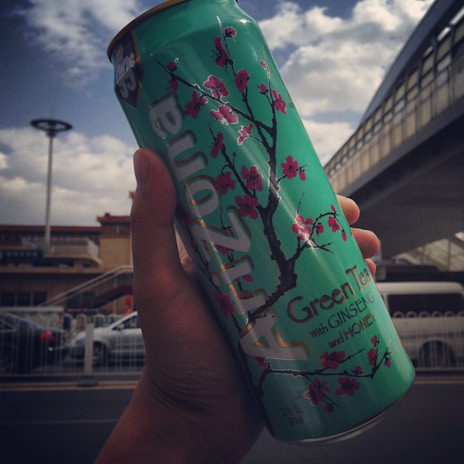 A person is holding a large can of Arizona Green Tea with Ginseng and Honey. The can features a light green color with a design of blooming pink cherry blossoms. It is held against a backdrop of an urban setting with a cloudy sky, a pedestrian bridge, and some buildings in the distance.
