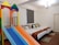 A brightly lit room contains two beds with colorful pillows and brown runners. Adjacent to the beds is a vibrant children's play structure with a colorful slide that includes shades of orange, blue, yellow, green, and red. The room has wooden flooring and a simple chandelier hanging from the ceiling.