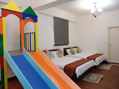A brightly lit room contains two beds with colorful pillows and brown runners. Adjacent to the beds is a vibrant children's play structure with a colorful slide that includes shades of orange, blue, yellow, green, and red. The room has wooden flooring and a simple chandelier hanging from the ceiling.