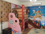 A colorful, whimsical room filled with vibrant decorations. There is a play structure resembling a gingerbread house, adorned with candy-themed designs, including lollipops and gumdrops. The walls are decorated with cheerful murals featuring stars, clouds, teddy bears, and a castle, all in bright, playful colors. The overall atmosphere is child-friendly, with a fantasy theme.