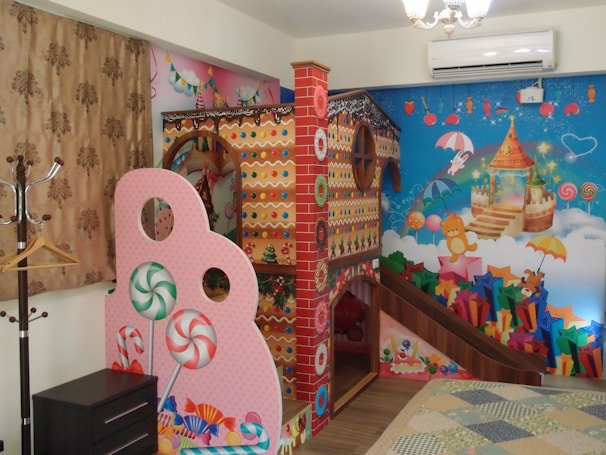 A colorful, whimsical room filled with vibrant decorations. There is a play structure resembling a gingerbread house, adorned with candy-themed designs, including lollipops and gumdrops. The walls are decorated with cheerful murals featuring stars, clouds, teddy bears, and a castle, all in bright, playful colors. The overall atmosphere is child-friendly, with a fantasy theme.