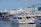 A marina filled with several white luxury yachts docked alongside a pier. In the background, there is industrial infrastructure, including a large crane labeled 'Wise 80 Ton.' The sky is clear with a few clouds visible, and the surrounding area is bustling with maritime activity.