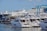 A marina filled with several white luxury yachts docked alongside a pier. In the background, there is industrial infrastructure, including a large crane labeled 'Wise 80 Ton.' The sky is clear with a few clouds visible, and the surrounding area is bustling with maritime activity.