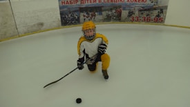 A young ice hockey player is kneeling on an ice rink, wearing yellow and black protective gear including a helmet, gloves, and skates. They are holding a hockey stick near a puck on the ice. A board with advertisements and images of other hockey players is visible in the background.