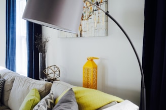 A modern interior living space featuring a large floor lamp arching over a corner of a soft white sofa adorned with gray and green cushions. Nearby, a bright yellow decorative lantern sits on the floor, complemented by a geometric metal sculpture and a tall glass vase with dried branches on a small table. Blue curtains frame a window in the background, and a piece of abstract art hangs on the wall.