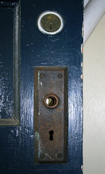 A rusty metal door lock and keyhole plate on a painted wooden door. The lock is old and has visible signs of wear and corrosion. Above the plate, there is a brass deadbolt lock.