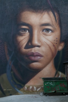 A detailed mural of a young person's face with realistic features and intricate details occupies a large portion of the wall. The facial expression is thoughtful, and there are decorative marks on the cheeks. Below the mural, a green dumpster is present, which is partially covered with graffiti.