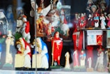 A collection of colorful religious figurines and statuettes on display, including representations of saints, angels, and other religious symbols. The statues are highly detailed, with some dressed in red and others in blue or white garments. A prominent figure in red is holding a scythe, and there is also a sign reading 'Poderoso Talisman Super Protector'.
