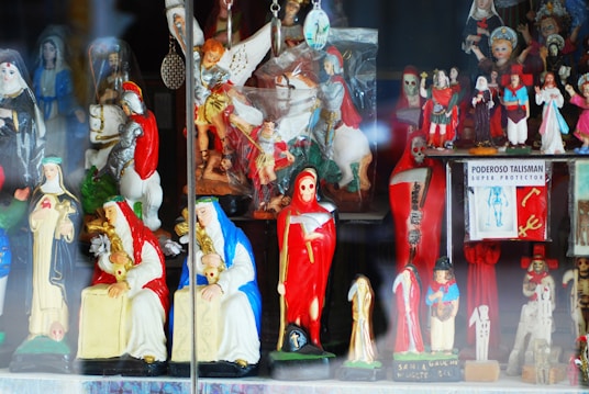 A collection of colorful religious figurines and statuettes on display, including representations of saints, angels, and other religious symbols. The statues are highly detailed, with some dressed in red and others in blue or white garments. A prominent figure in red is holding a scythe, and there is also a sign reading 'Poderoso Talisman Super Protector'.