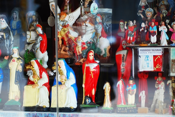 A collection of colorful religious figurines and statuettes on display, including representations of saints, angels, and other religious symbols. The statues are highly detailed, with some dressed in red and others in blue or white garments. A prominent figure in red is holding a scythe, and there is also a sign reading 'Poderoso Talisman Super Protector'.