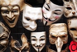 A collection of intricately designed masks with various expressions arranged against a dark background. Some masks have ornate decorations and distinct features such as curled mustaches and rosy cheeks, while others display exaggerated facial features.