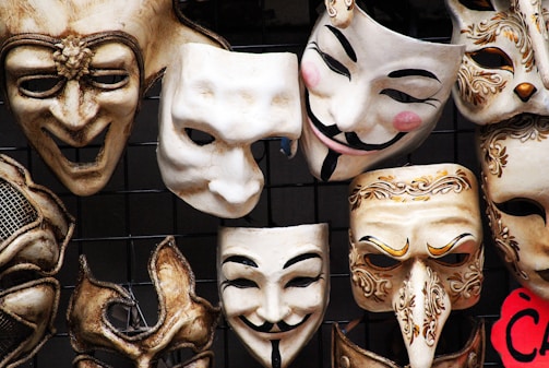 A collection of intricately designed masks with various expressions arranged against a dark background. Some masks have ornate decorations and distinct features such as curled mustaches and rosy cheeks, while others display exaggerated facial features.